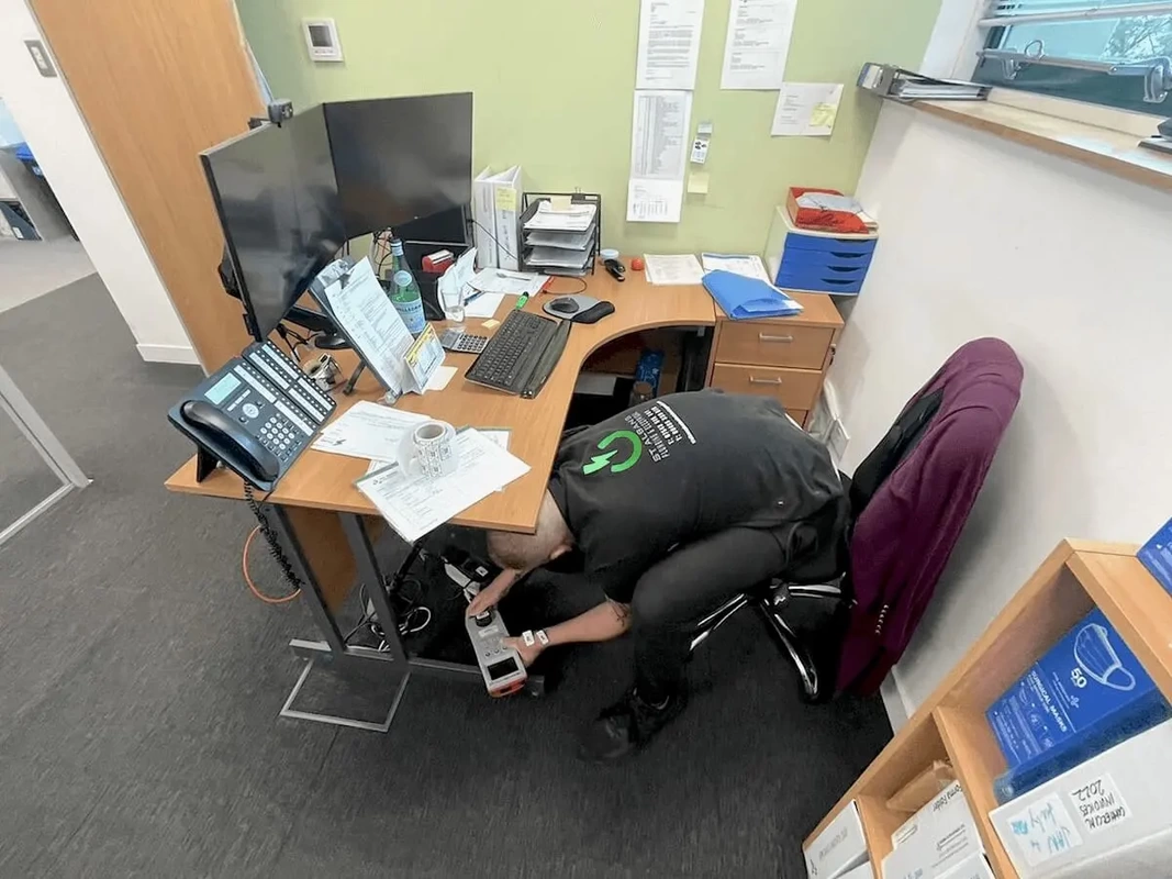 PAT Testing Watford Engineer PAT Testing electrical equipment, leaning down while sitting in a desk chair holding a pat tester machine